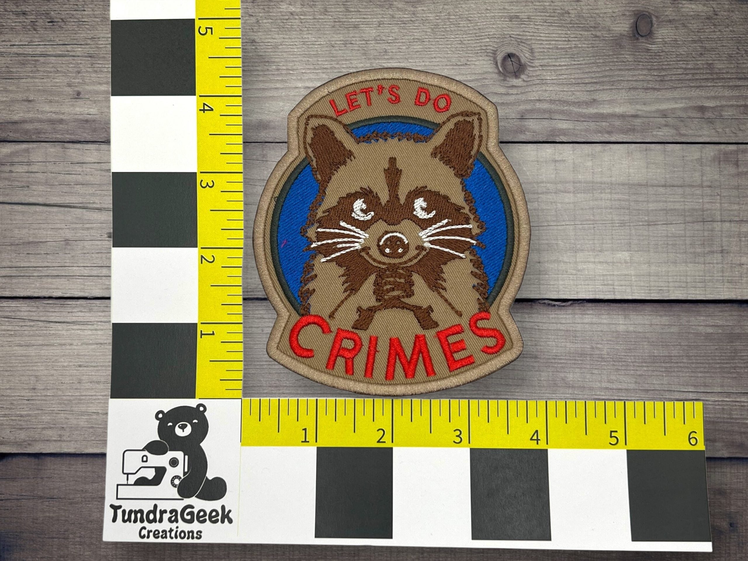 A patch featuring a raccoon with the text LETS DO CRIMES is placed on a wooden surface next to two measuring rulers and a TundraGeek Creations logo.