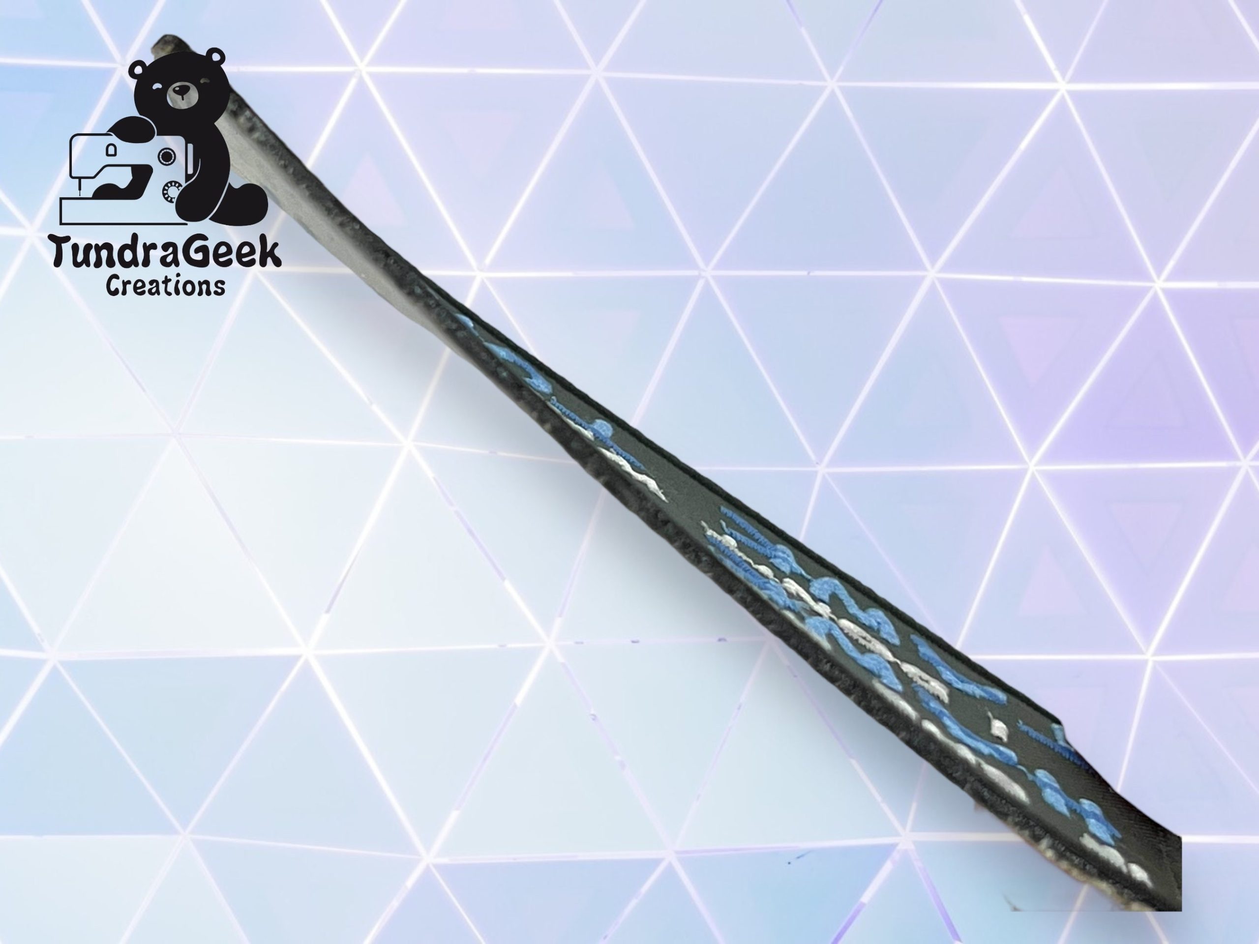 A handmade foam sword with blue and silver detailing lies on a geometric, light purple background. TundraGeek Creations logo appears in the top left corner.