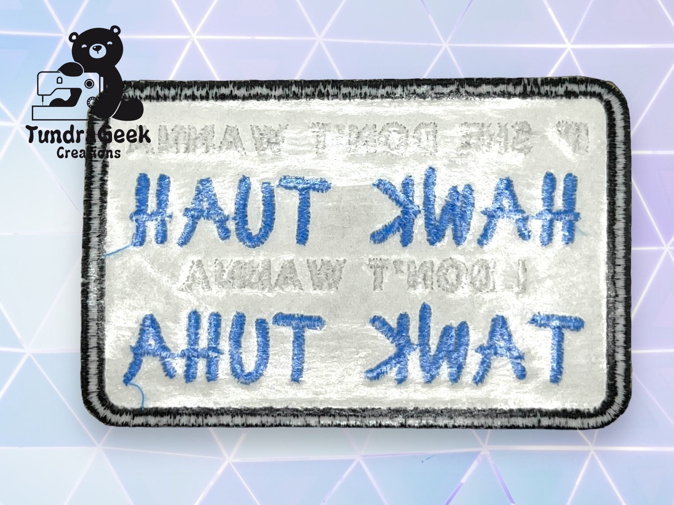 A rectangular embroidered patch viewed from the back, showing reversed text in blue and black thread; Tundra Geek Creations logo in the corner.
