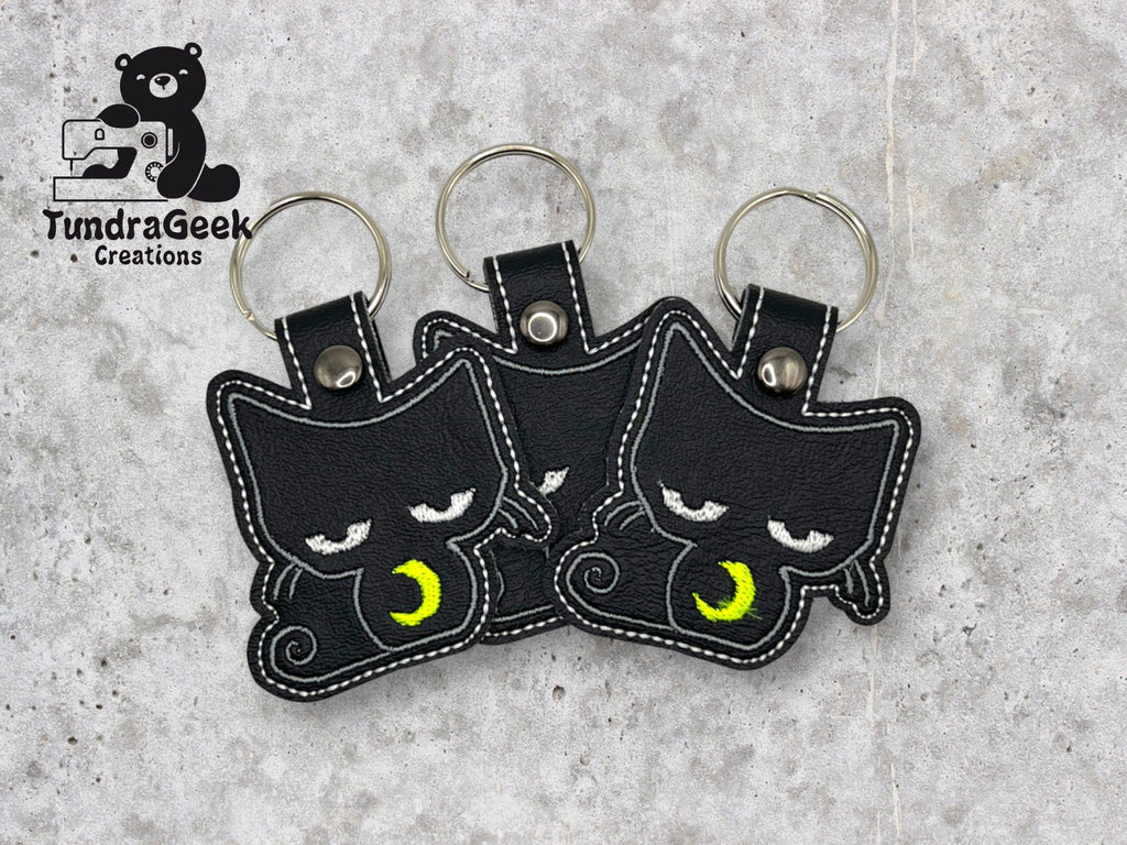 Three black cat-shaped keychains with embroidered yellow crescent moons on their chests are arranged on a gray surface. TundraGeek Creations logo is in the top left corner.