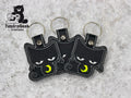Three black cat-shaped keychains with embroidered yellow crescent moons on their chests are arranged on a gray surface. TundraGeek Creations logo is in the top left corner.