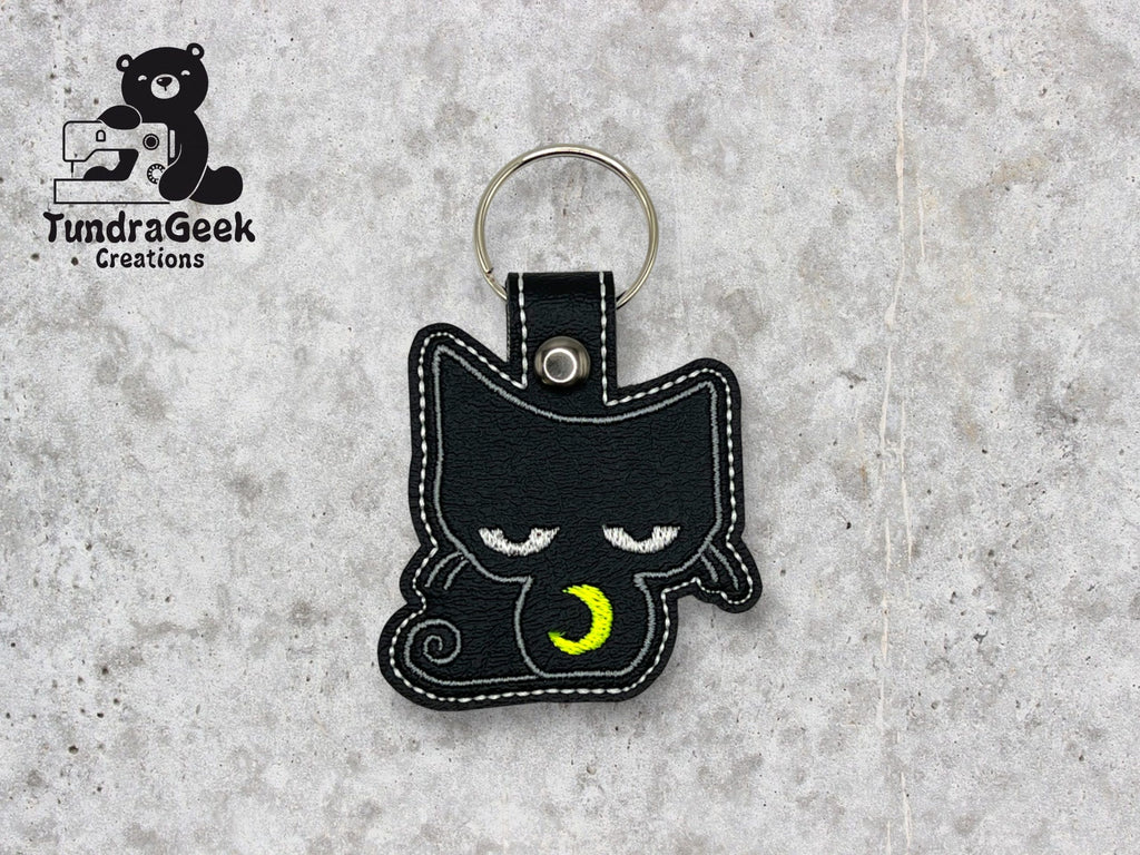 Black cat-shaped keychain with embroidered sleepy eyes and a yellow crescent moon, placed on a gray surface. TundraGeek Creations logo is visible in the top left corner.