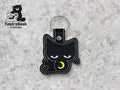 Black cat-shaped keychain with embroidered sleepy eyes and a yellow crescent moon, placed on a gray surface. TundraGeek Creations logo is visible in the top left corner.