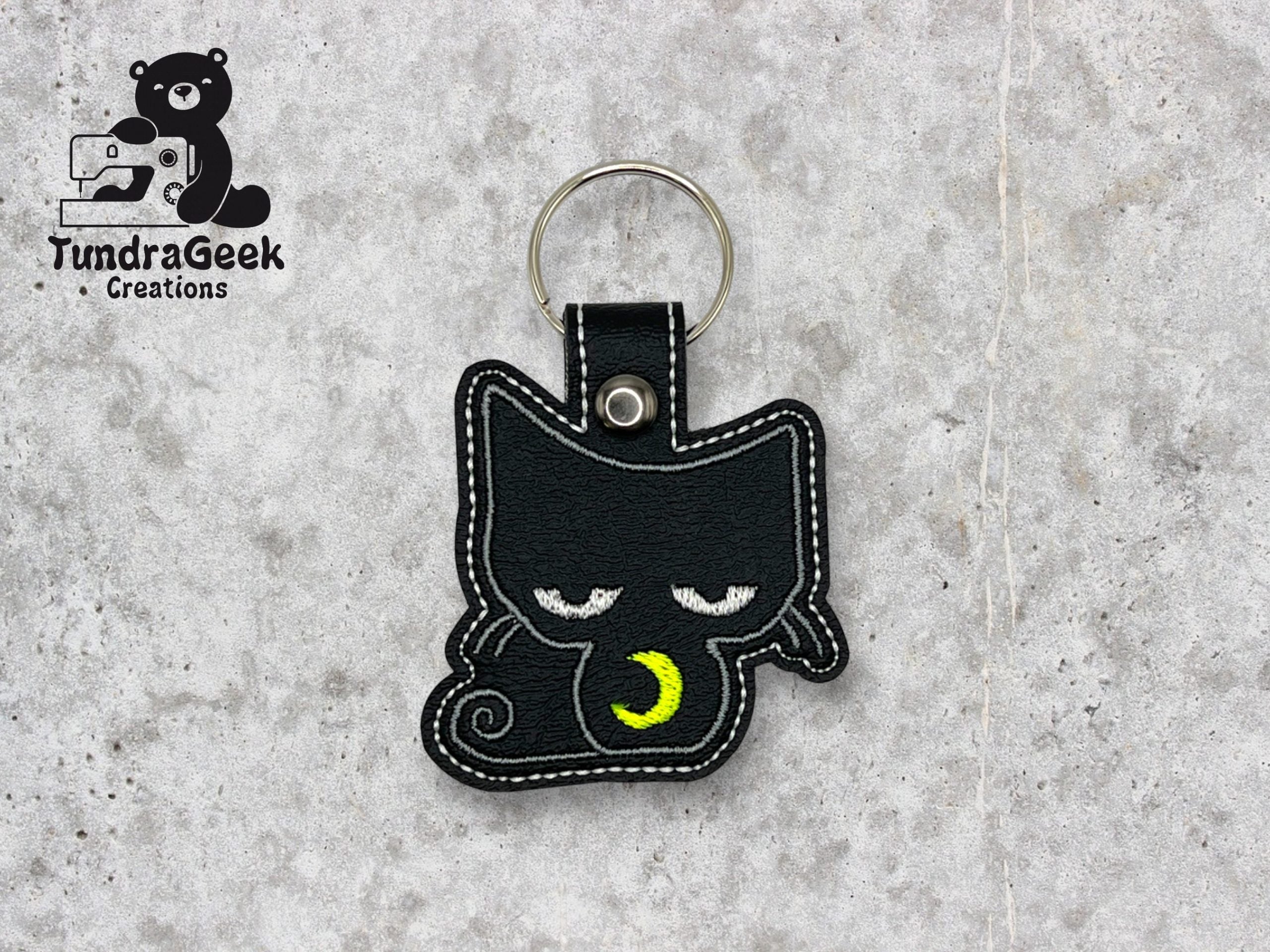 Black cat-shaped keychain with embroidered sleepy eyes and a yellow crescent moon, placed on a gray surface. TundraGeek Creations logo is visible in the top left corner.