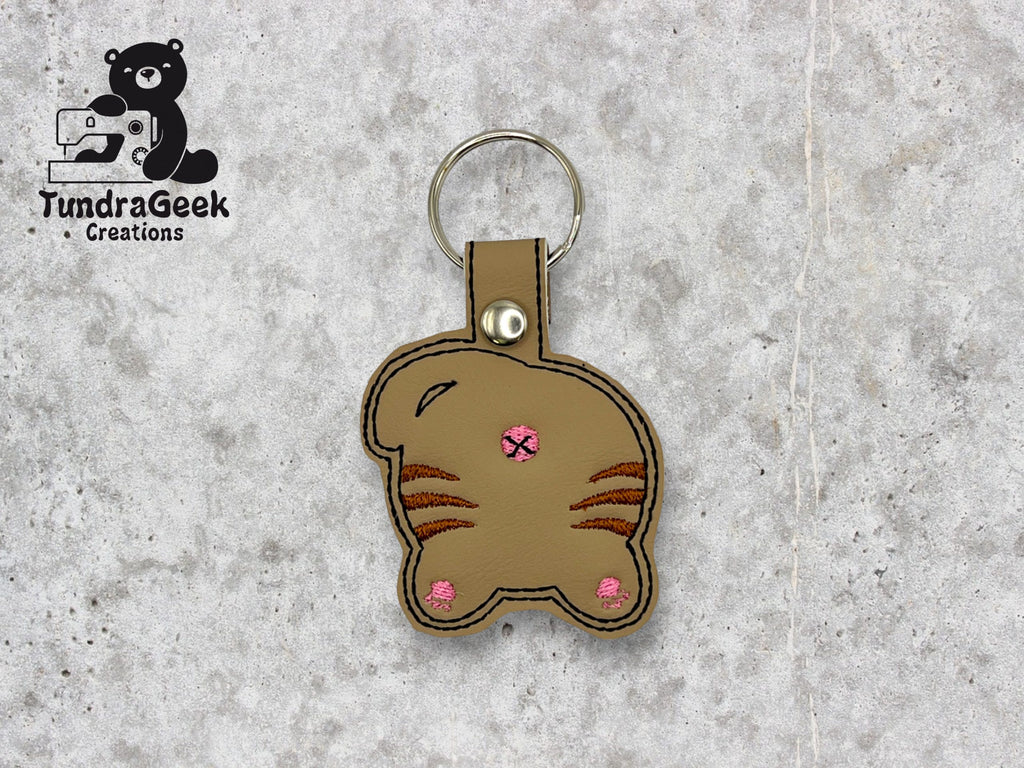 A keychain shaped like the back of a brown cat with pink embroidered details, displayed on a light stone background. TundraGeek Creations logo is in the top left corner.