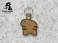 A keychain shaped like the back of a brown cat with pink embroidered details, displayed on a light stone background. TundraGeek Creations logo is in the top left corner.