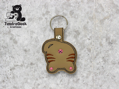 A keychain shaped like the back of a brown cat with pink embroidered details, displayed on a light stone background. TundraGeek Creations logo is in the top left corner.