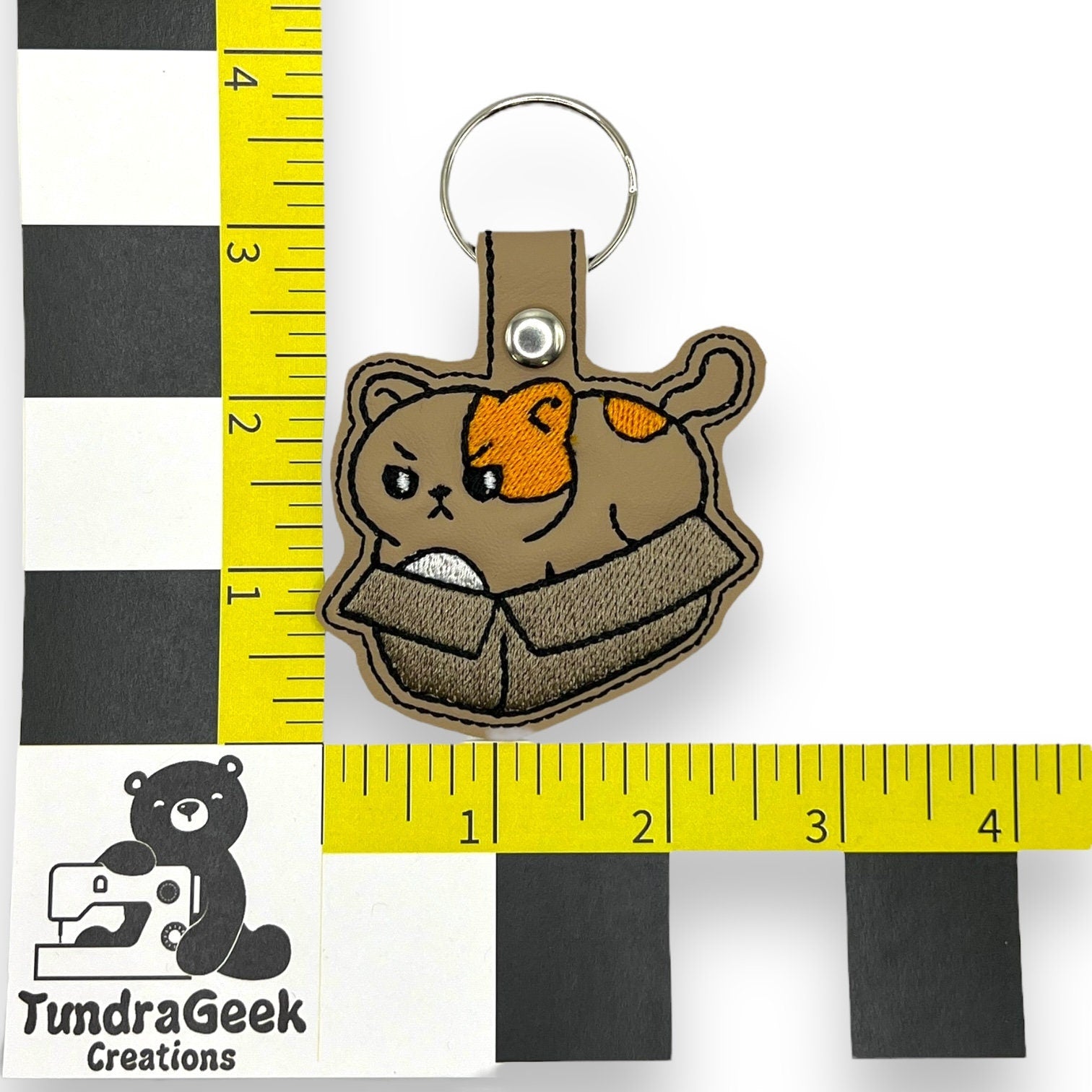 A cat keychain with an embroidered design of a brown and orange cat in a box, displayed alongside a measuring grid and TundraGeek Creations logo.