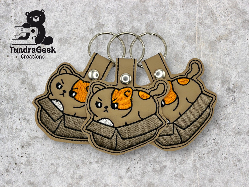 Three cat-in-box embroidered keychains displayed on a gray surface, with a black TundraGeek Creations logo in the top left corner.
