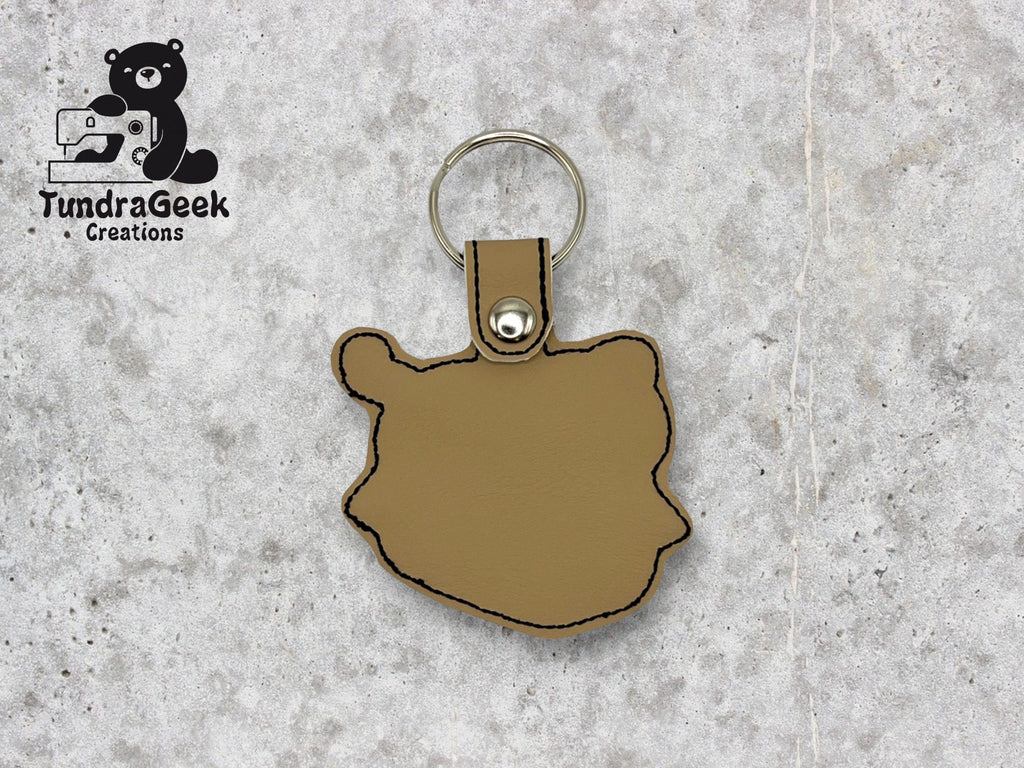A tan leather keychain shaped like the outline of Alaska with black stitching, a metal snap, and a key ring. TundraGeek Creations logo is in the top left corner.