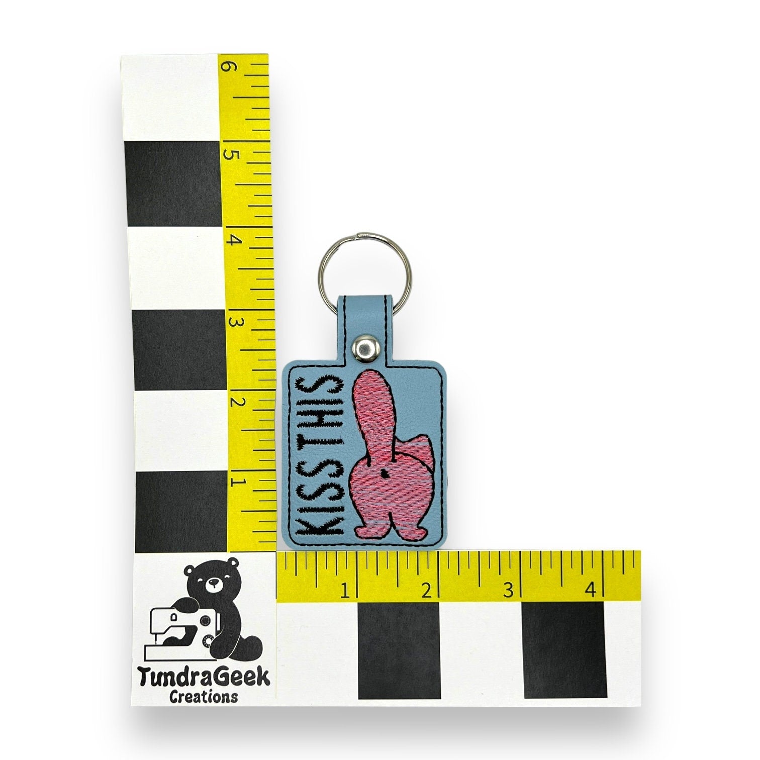 A blue keychain with a pink cartoon butt and KISS THIS text, placed on a measurement grid with TundraGeek Creations logo in the corner.