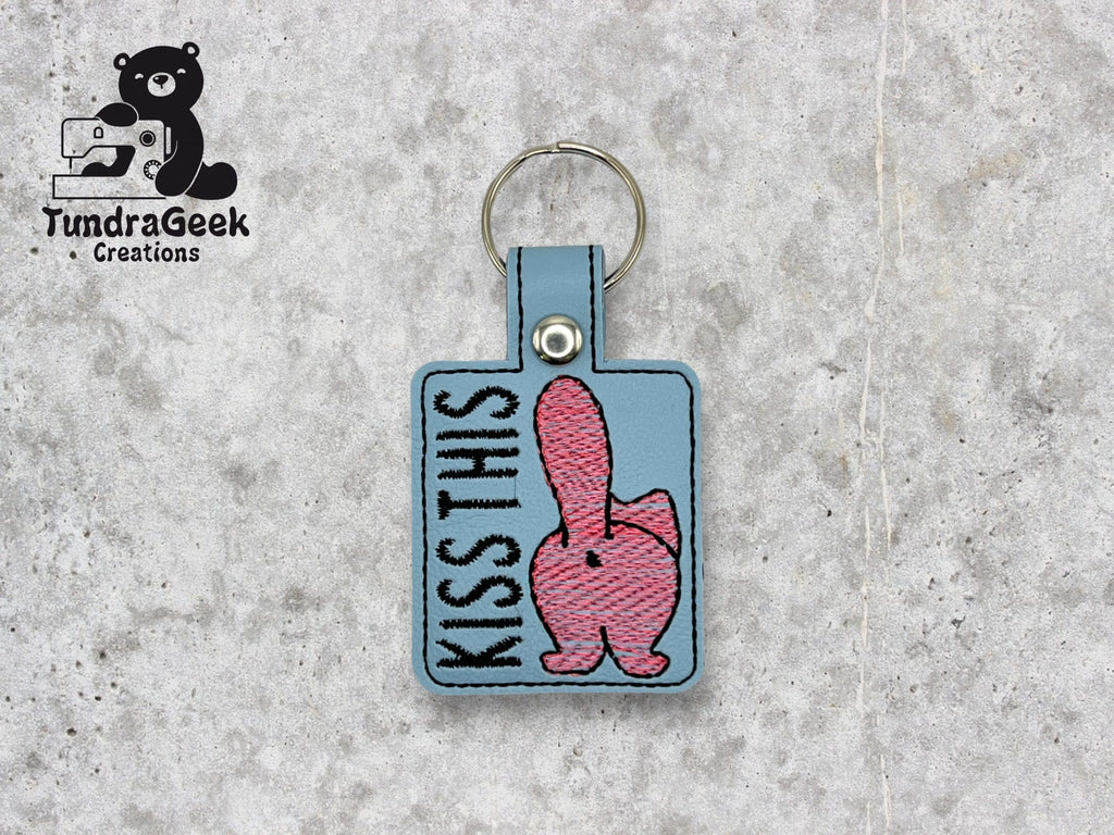 Embroidered keychain with a cartoon butt and the words KISS THIS on a gray background, with TundraGeek Creations logo in the top left corner.