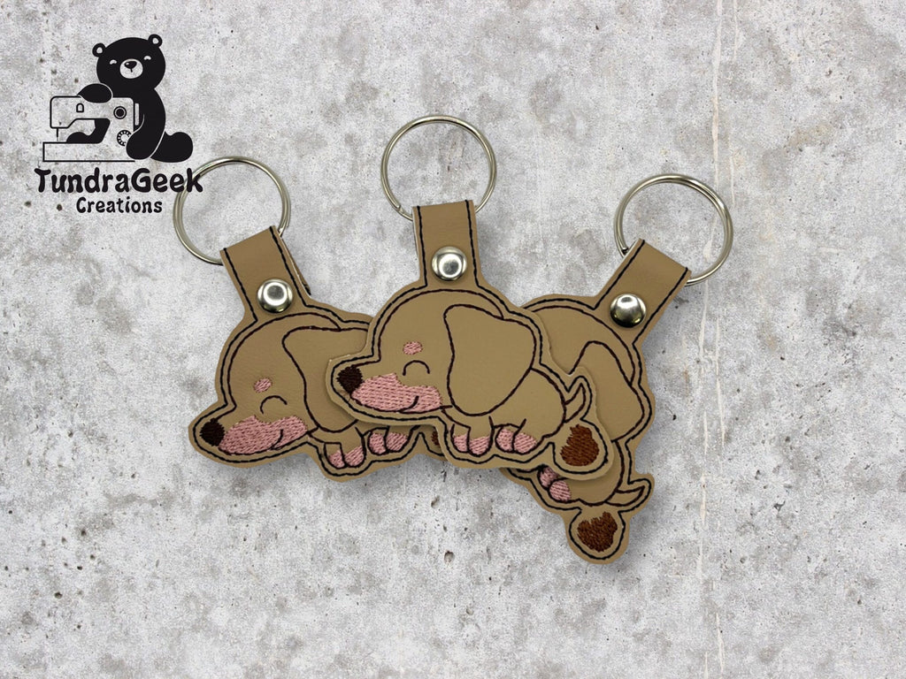 Three identical brown embroidered keychains shaped like sleeping dachshund puppies are displayed on a textured surface. The TundraGeek Creations logo appears in the top left corner.
