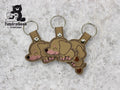 Three identical brown embroidered keychains shaped like sleeping dachshund puppies are displayed on a textured surface. The TundraGeek Creations logo appears in the top left corner.