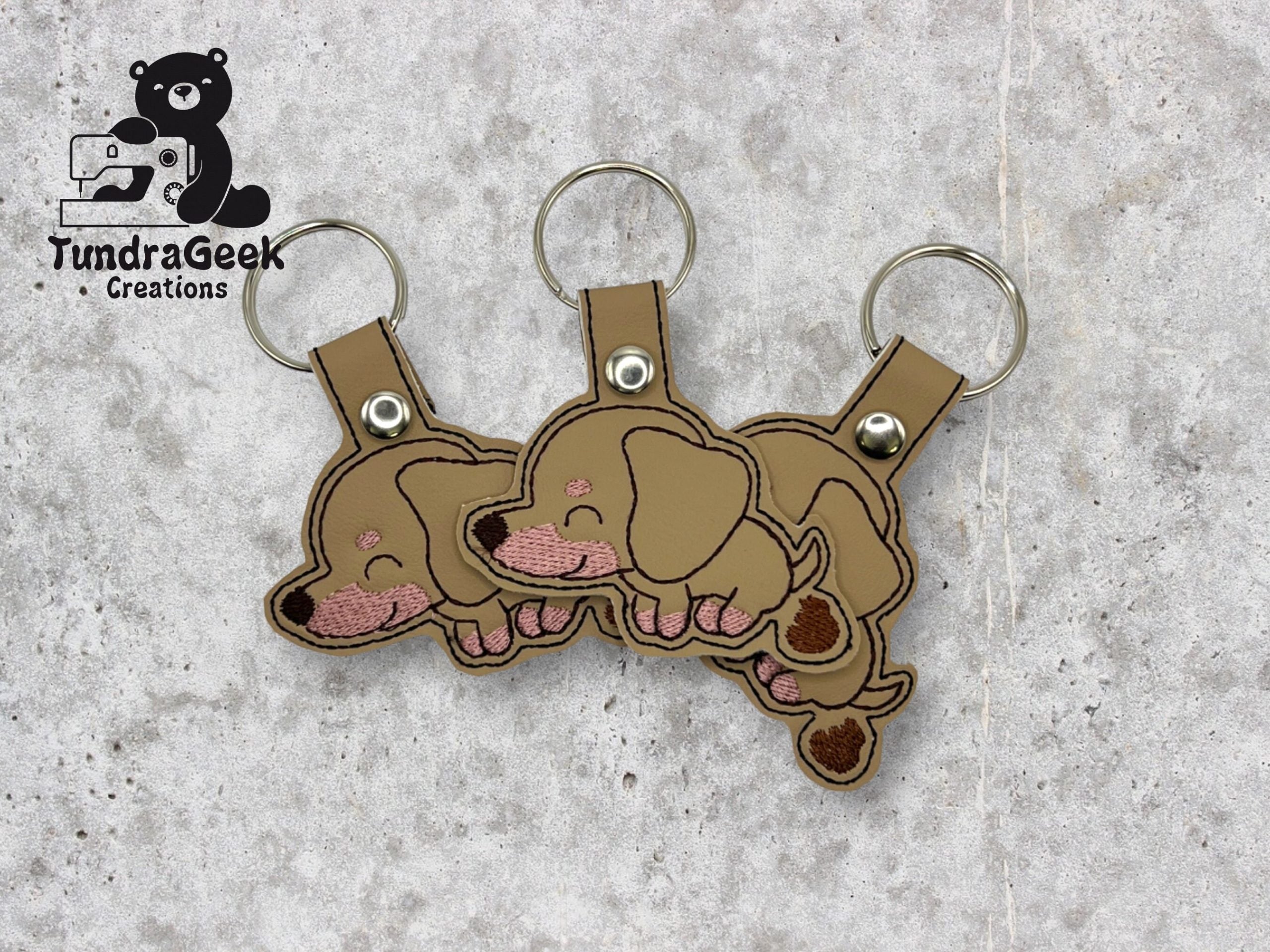 Three identical brown embroidered keychains shaped like sleeping dachshund puppies are displayed on a textured surface. The TundraGeek Creations logo appears in the top left corner.