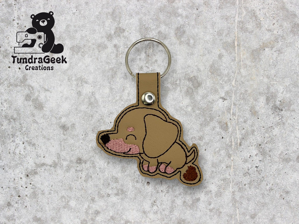 Tan and brown embroidered dachshund dog keychain with a snap closure on a light gray textured background. TundraGeek Creations logo is visible in the top left corner.