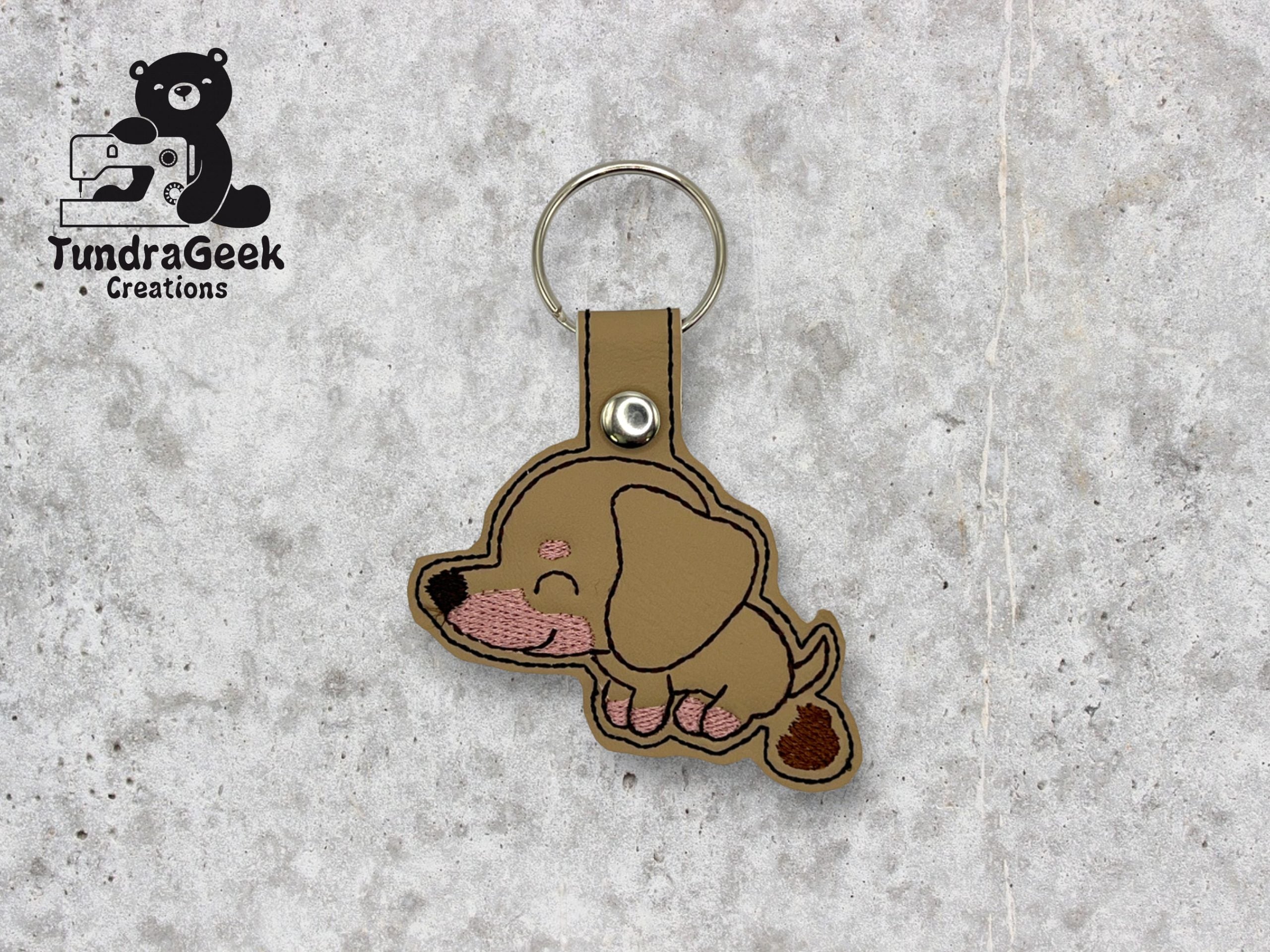Tan and brown embroidered dachshund dog keychain with a snap closure on a light gray textured background. TundraGeek Creations logo is visible in the top left corner.
