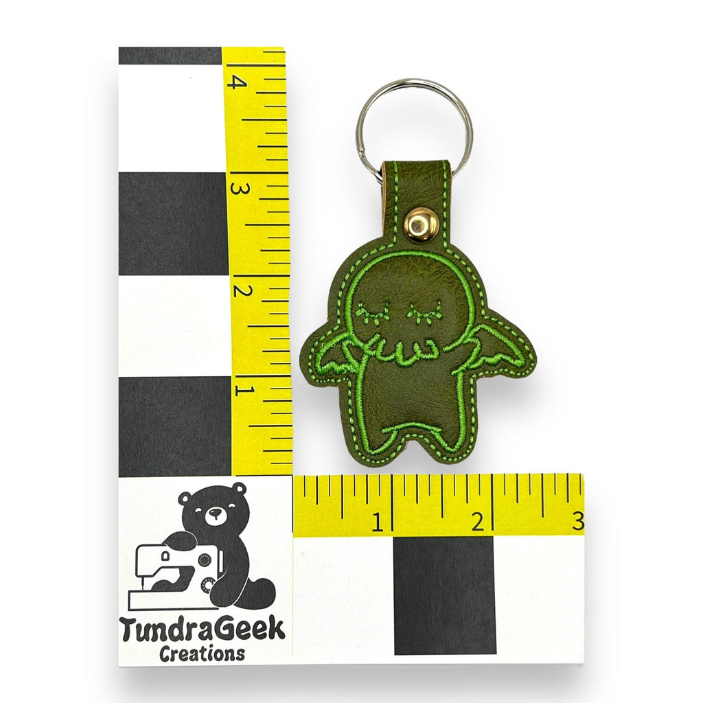 A small green leather keychain shaped like a cartoon creature is placed next to a ruler showing it is about 3 inches tall. A logo with a bear and text TundraGeek Creations is visible.