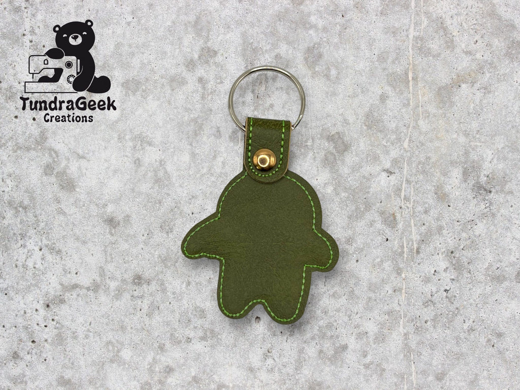 Green leather keychain in the shape of a simplified gingerbread figure with a metal ring, displayed on a gray surface. TundraGeek Creations logo is in the top left corner.