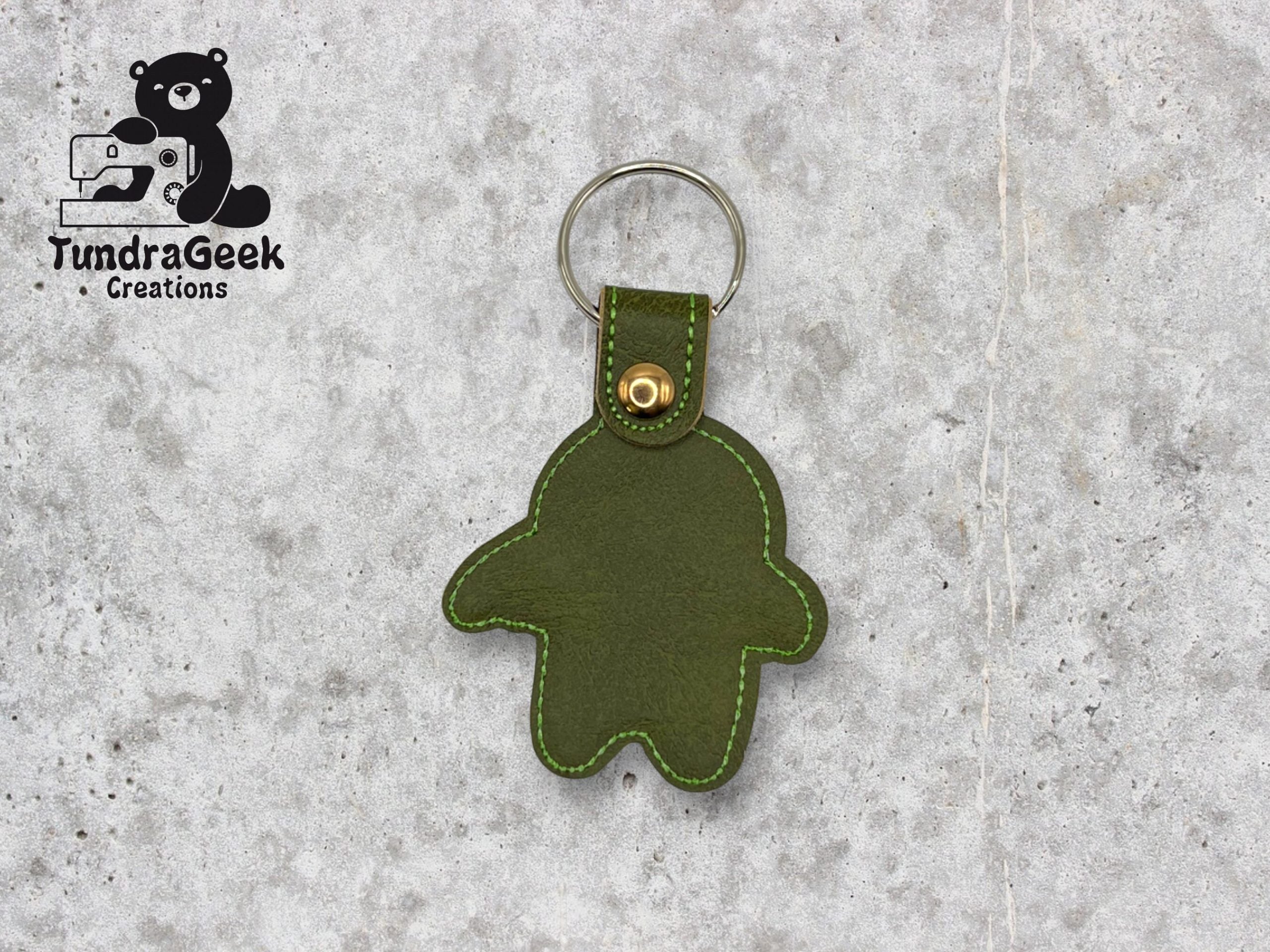 Green leather keychain in the shape of a simplified gingerbread figure with a metal ring, displayed on a gray surface. TundraGeek Creations logo is in the top left corner.