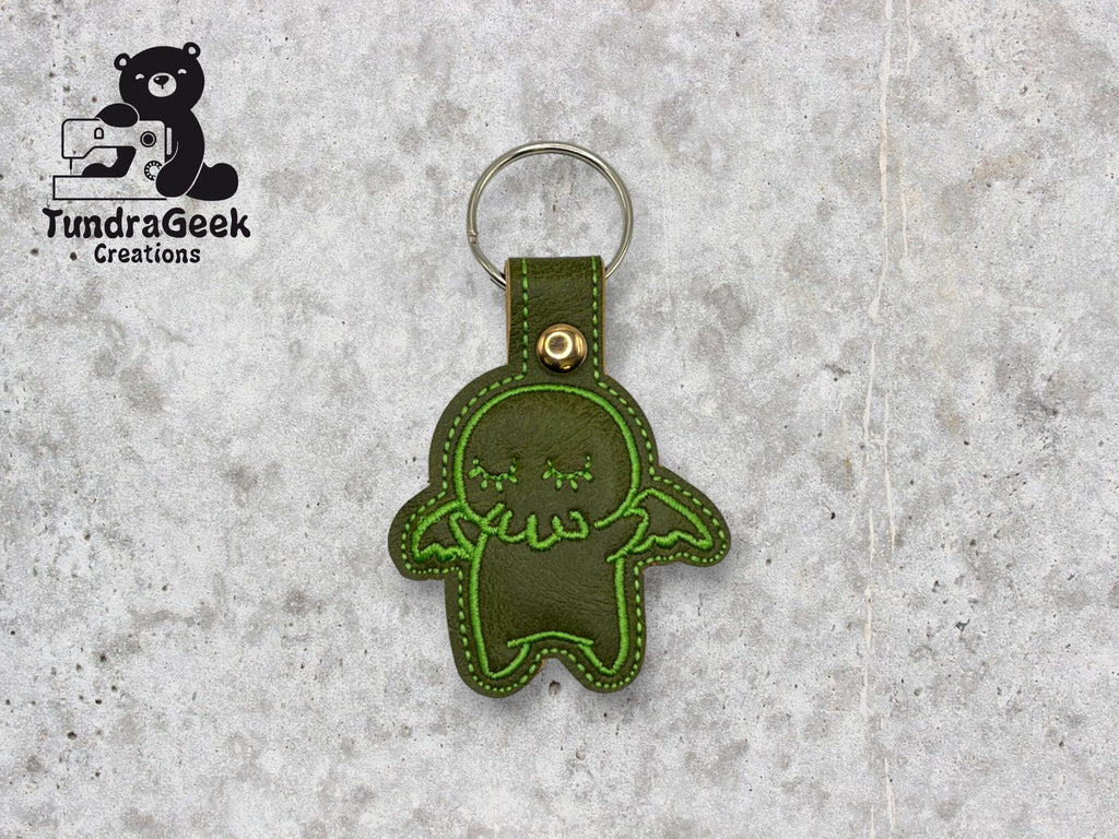 A green stitched keychain shaped like a cartoon creature with wings, displayed on a gray surface. The TundraGeek Creations logo is in the top left corner.