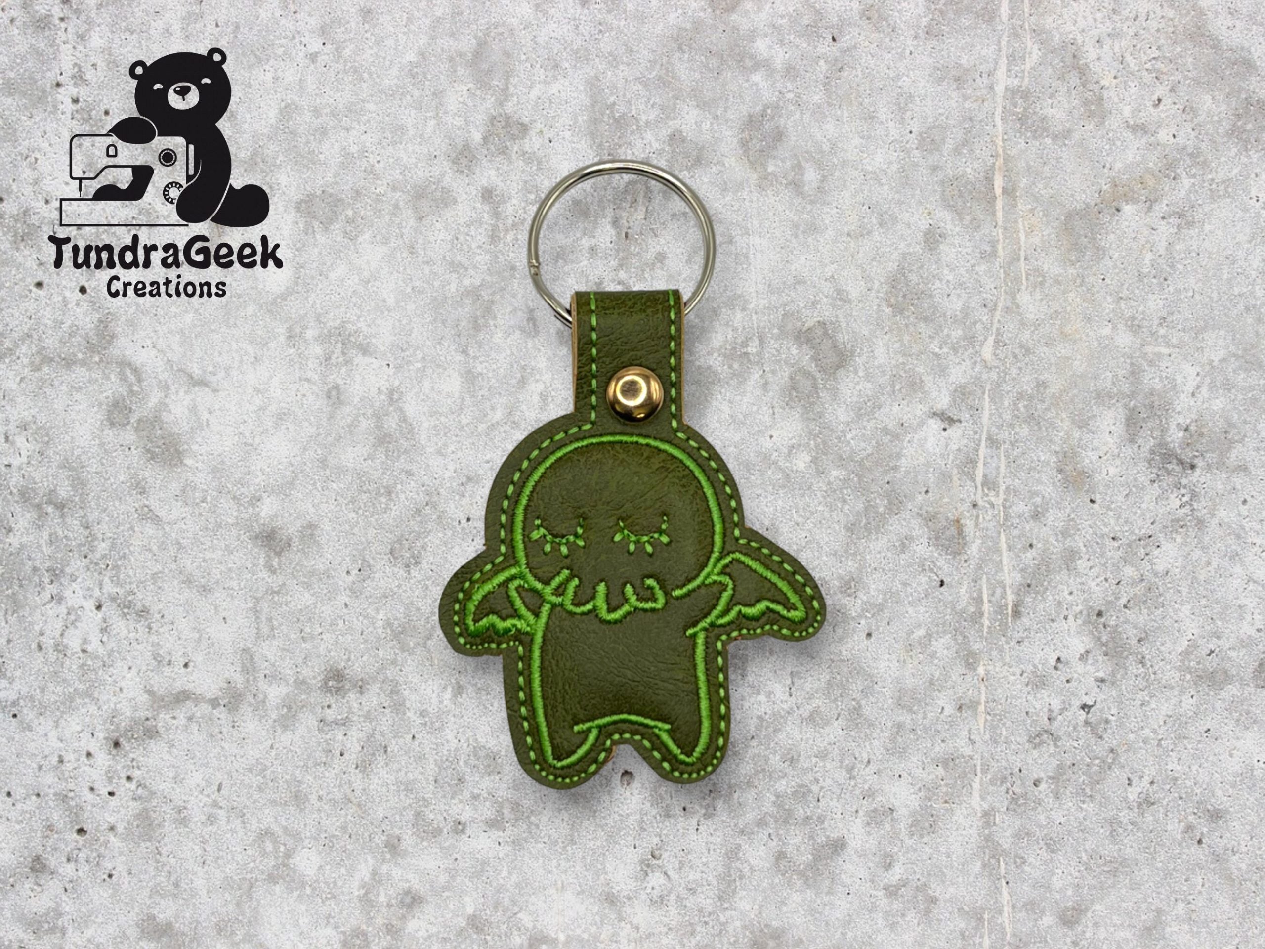 A green stitched keychain shaped like a cartoon creature with wings, displayed on a gray surface. The TundraGeek Creations logo is in the top left corner.