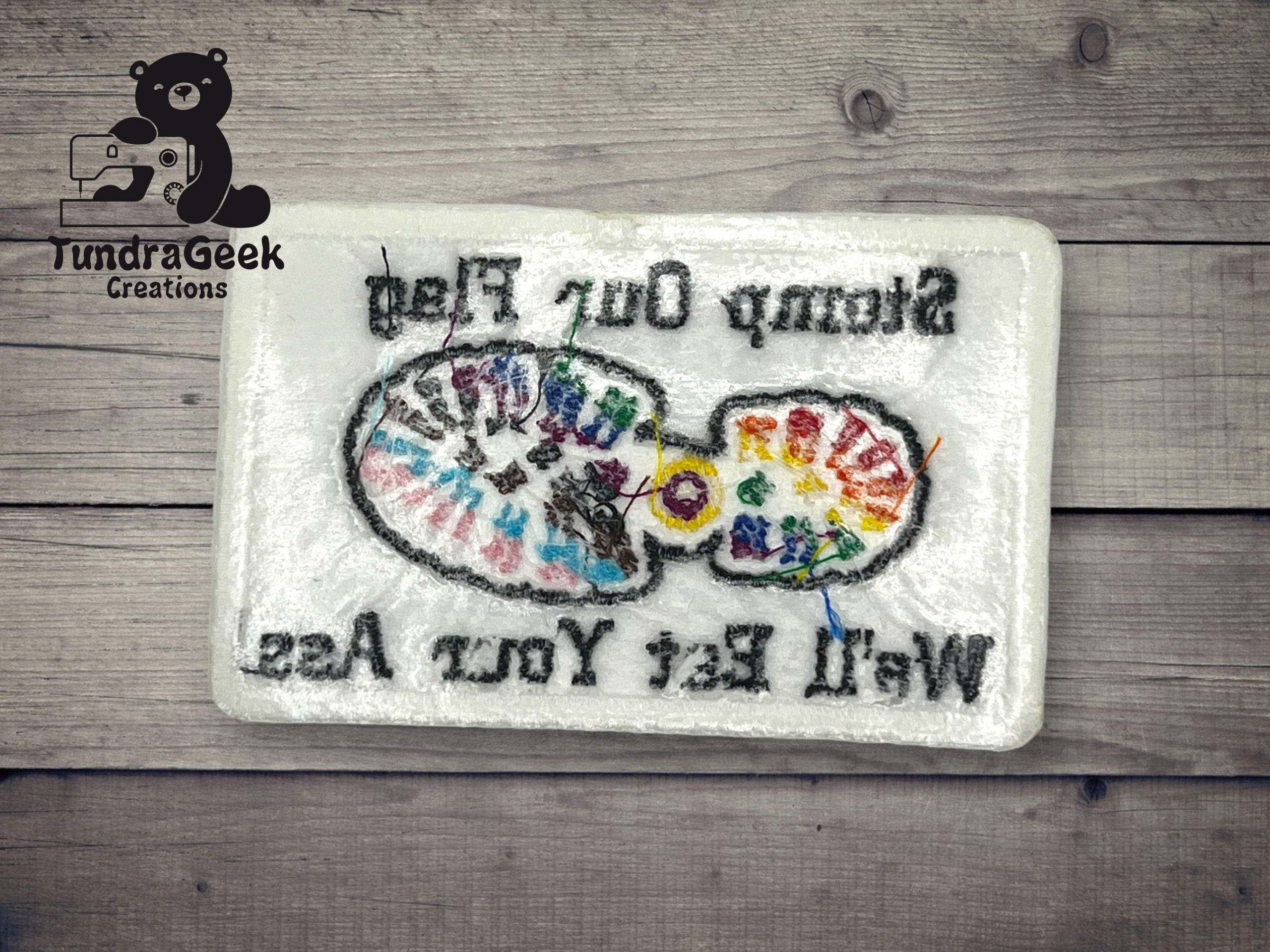 A rectangular felted mat with a colorful painters palette design and the text Stamp Out Rag, Wet Felting, You Ass in black lettering, on a wooden surface.