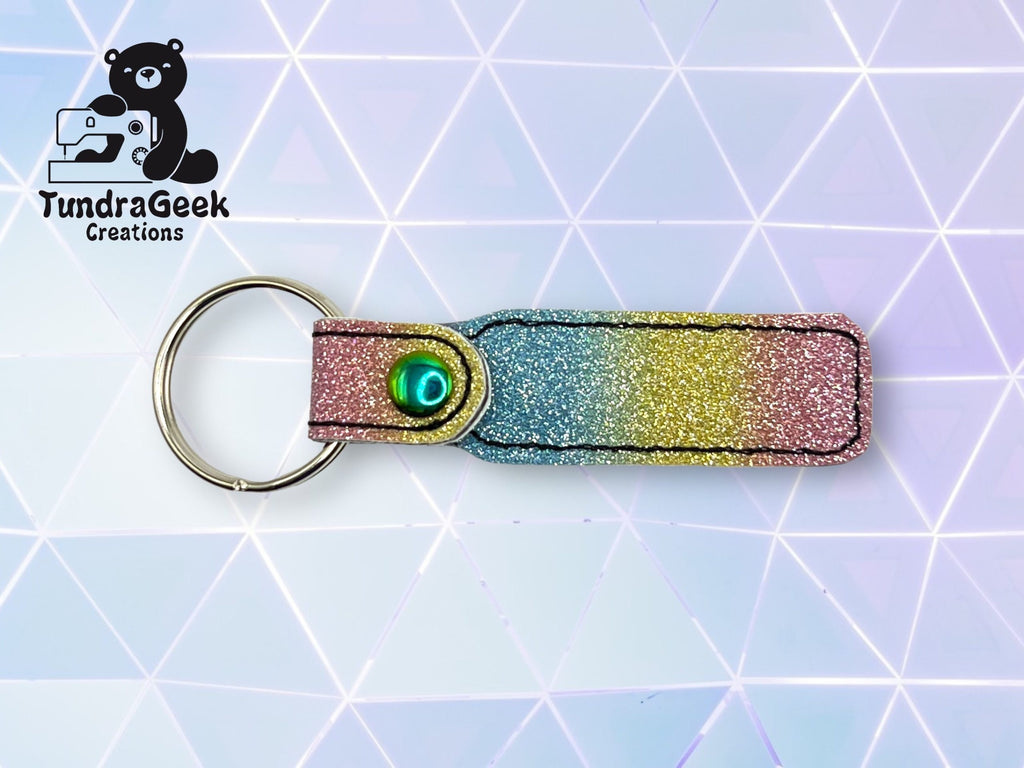 A glittery, multicolored keychain with a metal ring and a shiny green rivet, shown on a geometric pastel background. TundraGeek Creations logo in the top left corner.