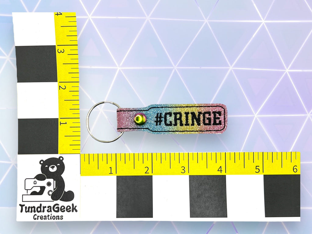 A keychain labeled #CRINGE is placed on a surface with geometric patterns, next to two yellow rulers showing its length is about 3 inches.