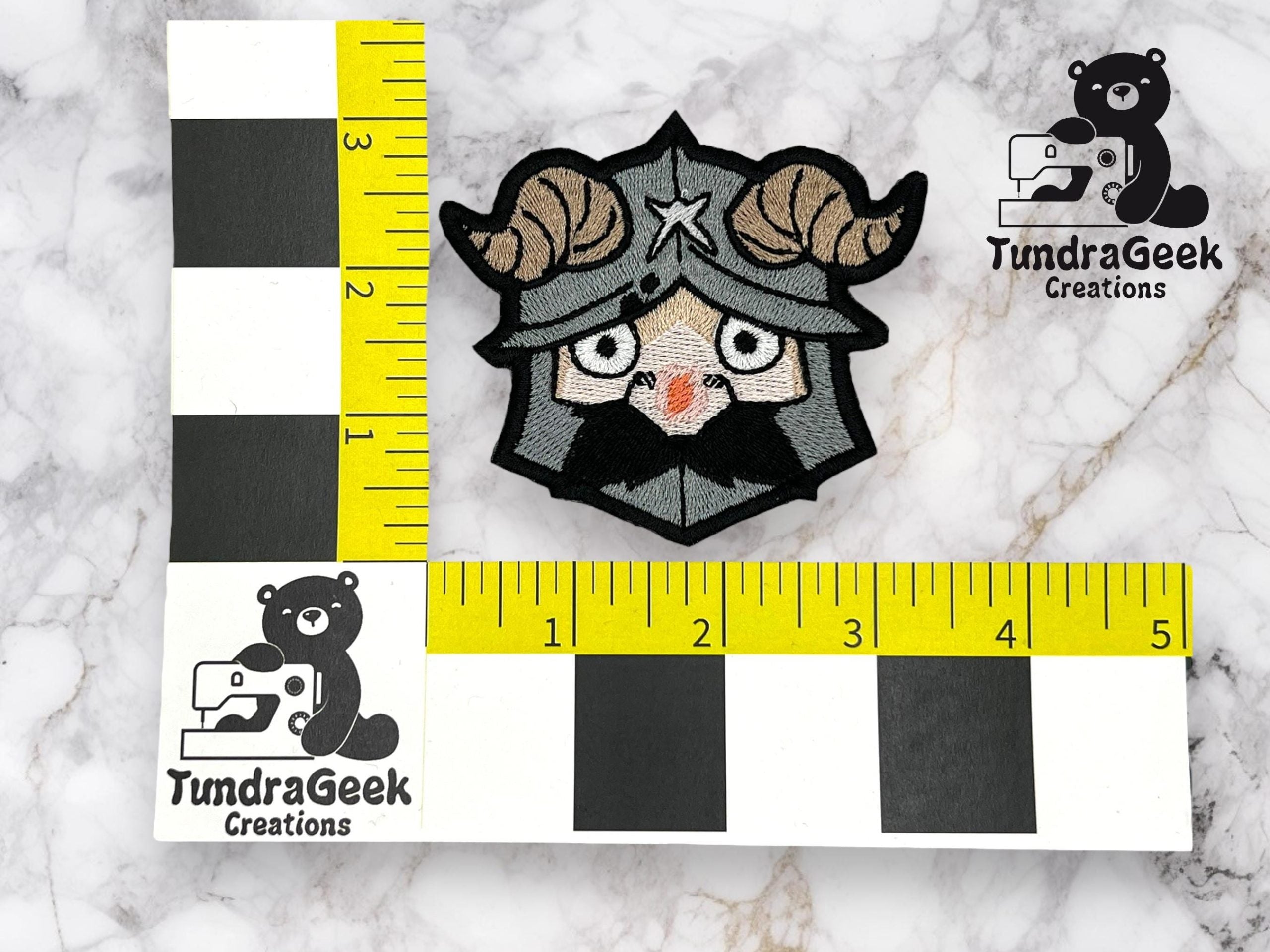 An embroidered patch of a horned helmeted character is shown on a marble surface, measured against yellow rulers marked with TundraGeek Creations logos.