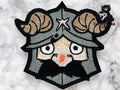 An embroidered patch depicts a cartoonish Viking with ram horns on a helmet, wide eyes, and a black beard. Marble surface background.