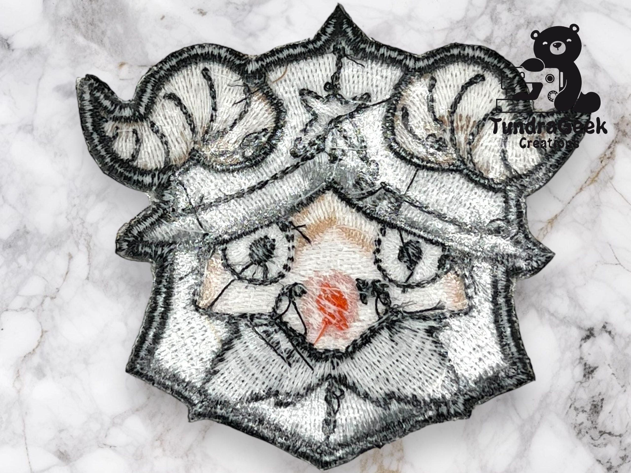 Embroidered patch depicting a stylized face with ram horns and a red nose, placed on a marble surface. The logo “Tundrasek Creations” with a bear is in the top right corner.