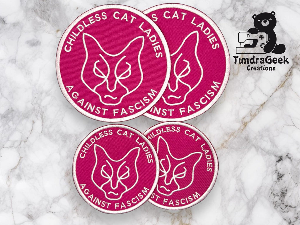Four magenta patches with a cat face outline and the words CHILDLESS CAT LADIES AGAINST FASCISM alongside the TundraGeek Creations logo on a marble background.