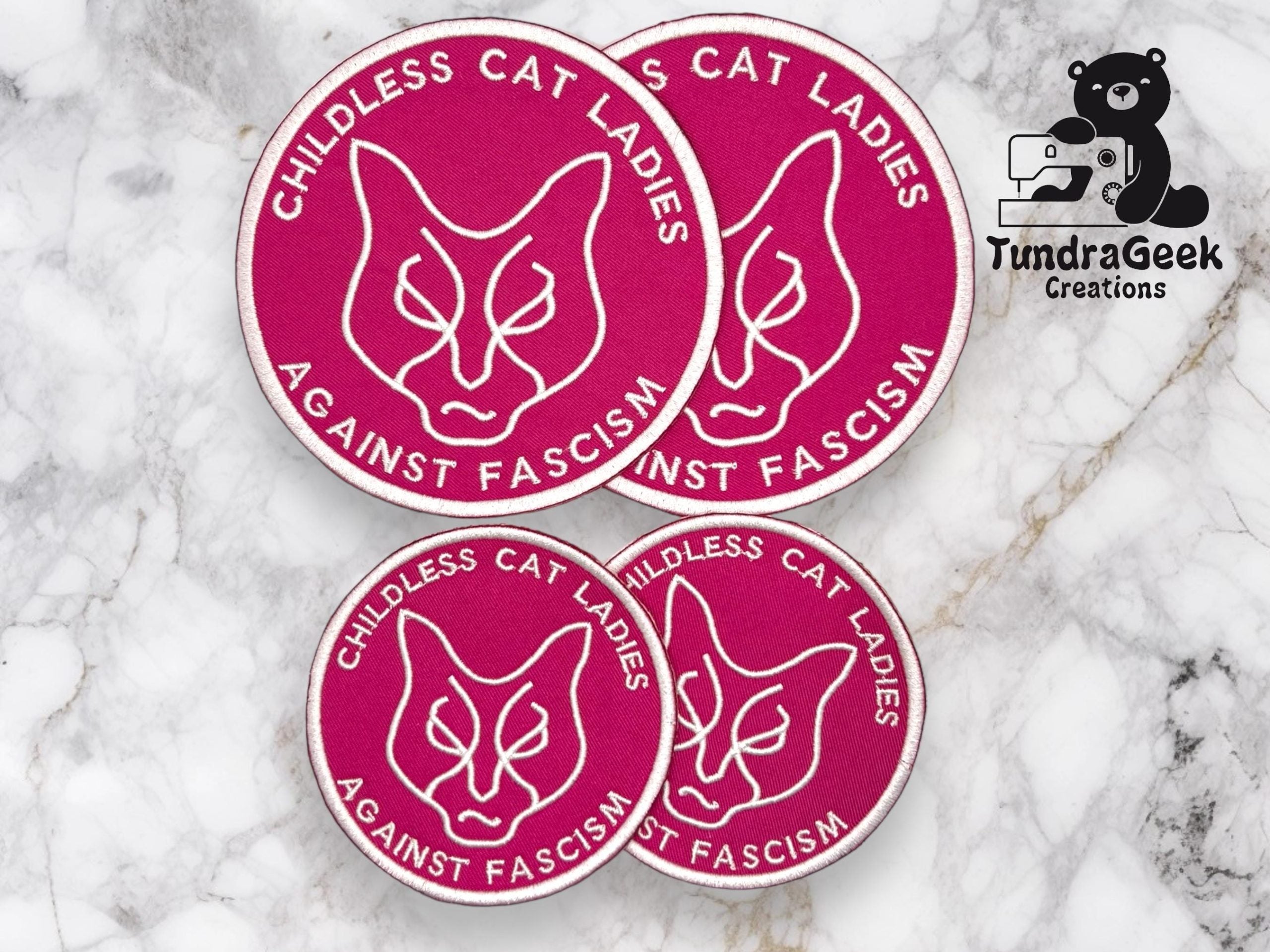 Four magenta patches with a cat face outline and the words CHILDLESS CAT LADIES AGAINST FASCISM alongside the TundraGeek Creations logo on a marble background.