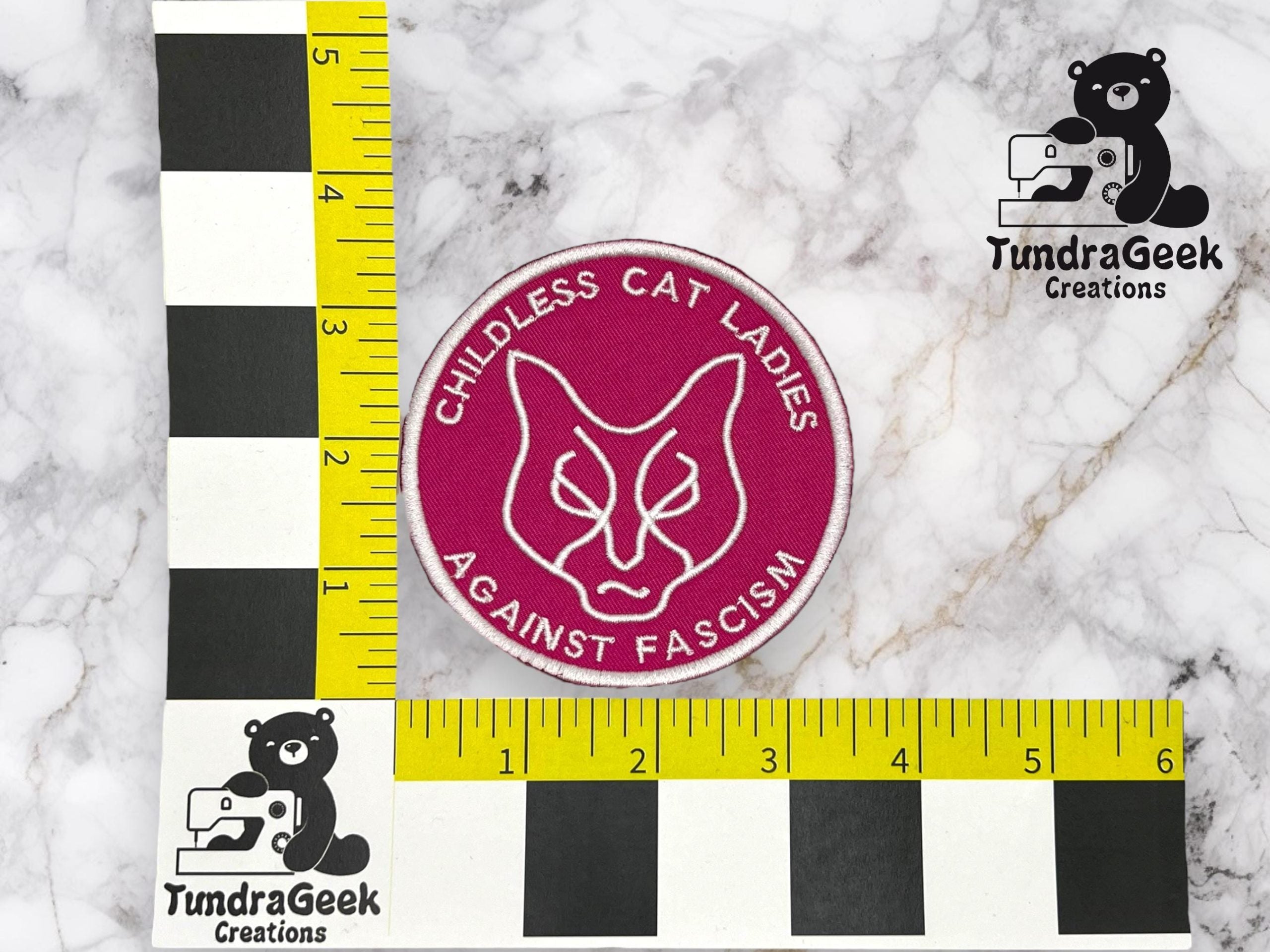A pink circular patch reads CHILDLESS CAT LADIES AGAINST FASCISM around a minimalist cat face; rulers show a diameter of about 3 inches. TundraGeek Creations logos are in the corners.