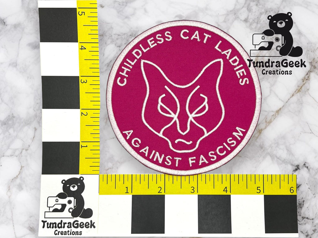 A round patch with a cat face and the text Childless Cat Ladies Against Fascism on a pink background, placed next to measurement rulers for scale.