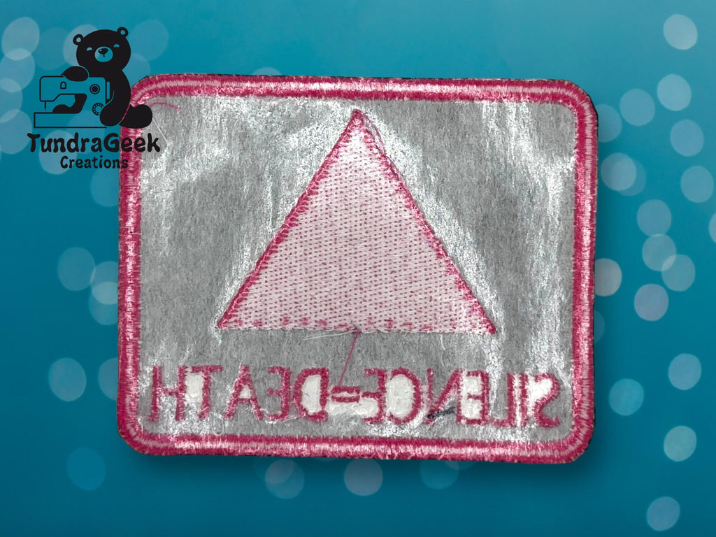 A rectangular embroidered patch with a pink triangle and the words SILENCE = DEATH in pink, shown in reverse. The background is silver with a pink border.