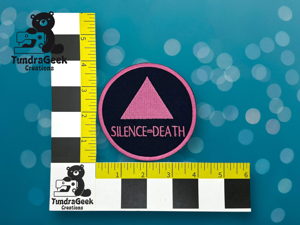 A circular patch with a pink triangle above the words Silence = Death is placed on a blue surface next to two yellow and black rulers showing size in inches.