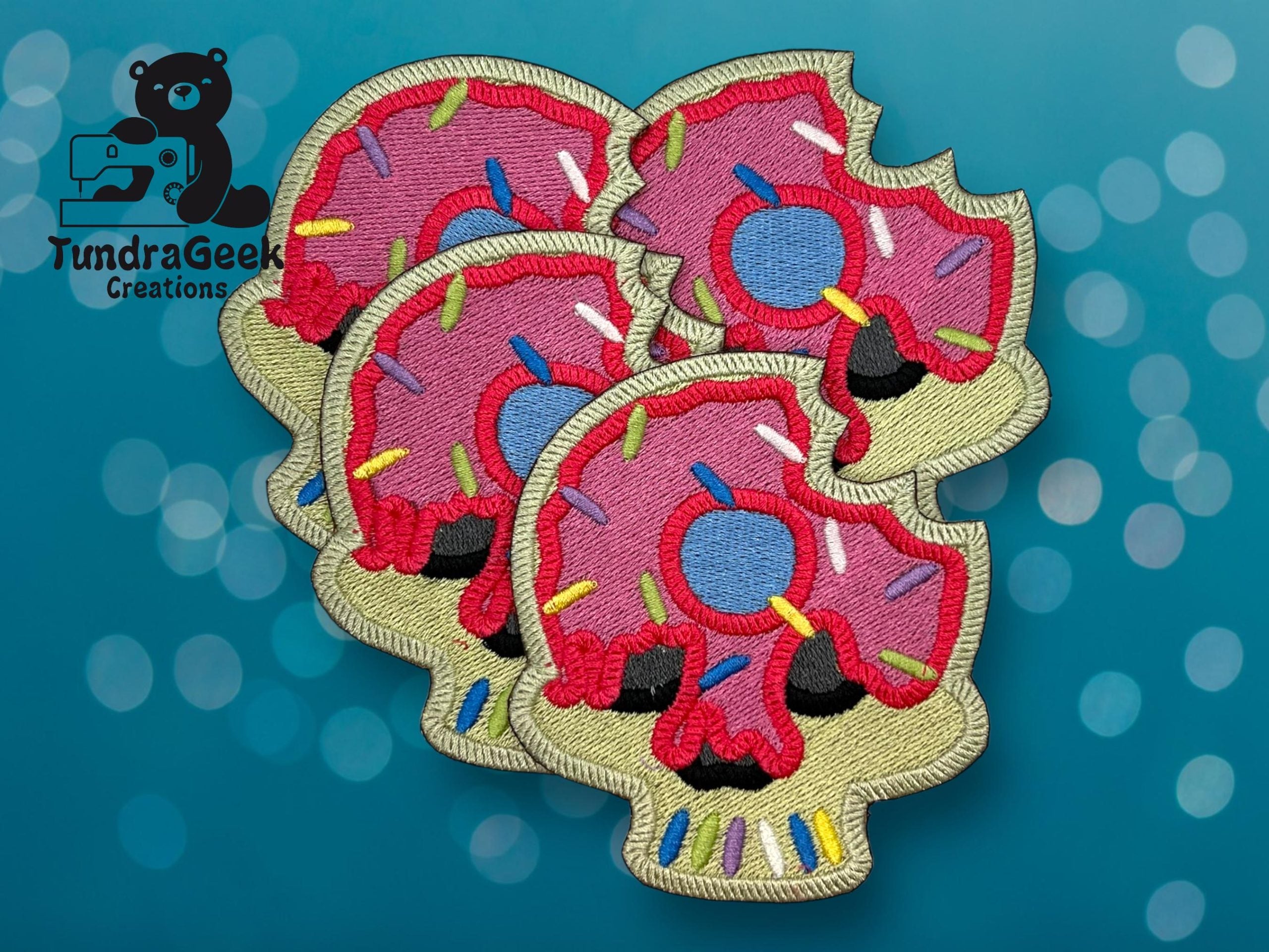 Four embroidered patches shaped like skulls with pink donut frosting and colorful sprinkles, shown on a blue background with the TundraGeek Creations logo in the corner.