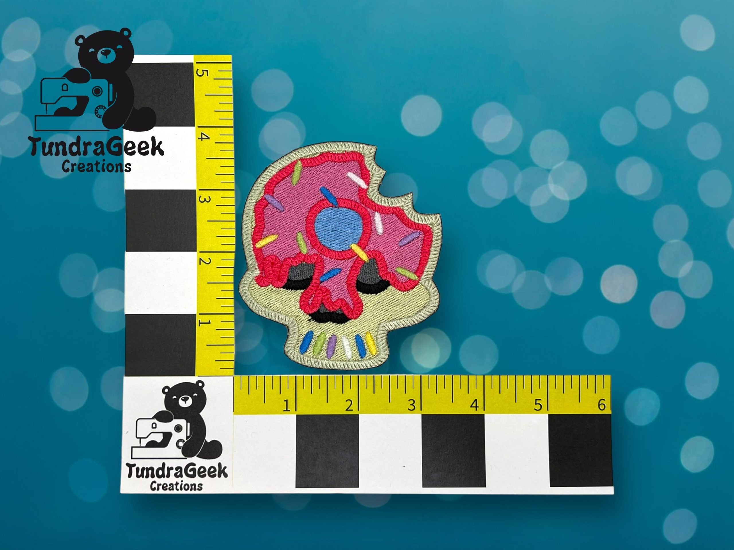 An embroidered patch shaped like a pink frosted donut skull with colorful sprinkles, placed next to a ruler showing a size of about 3 inches.