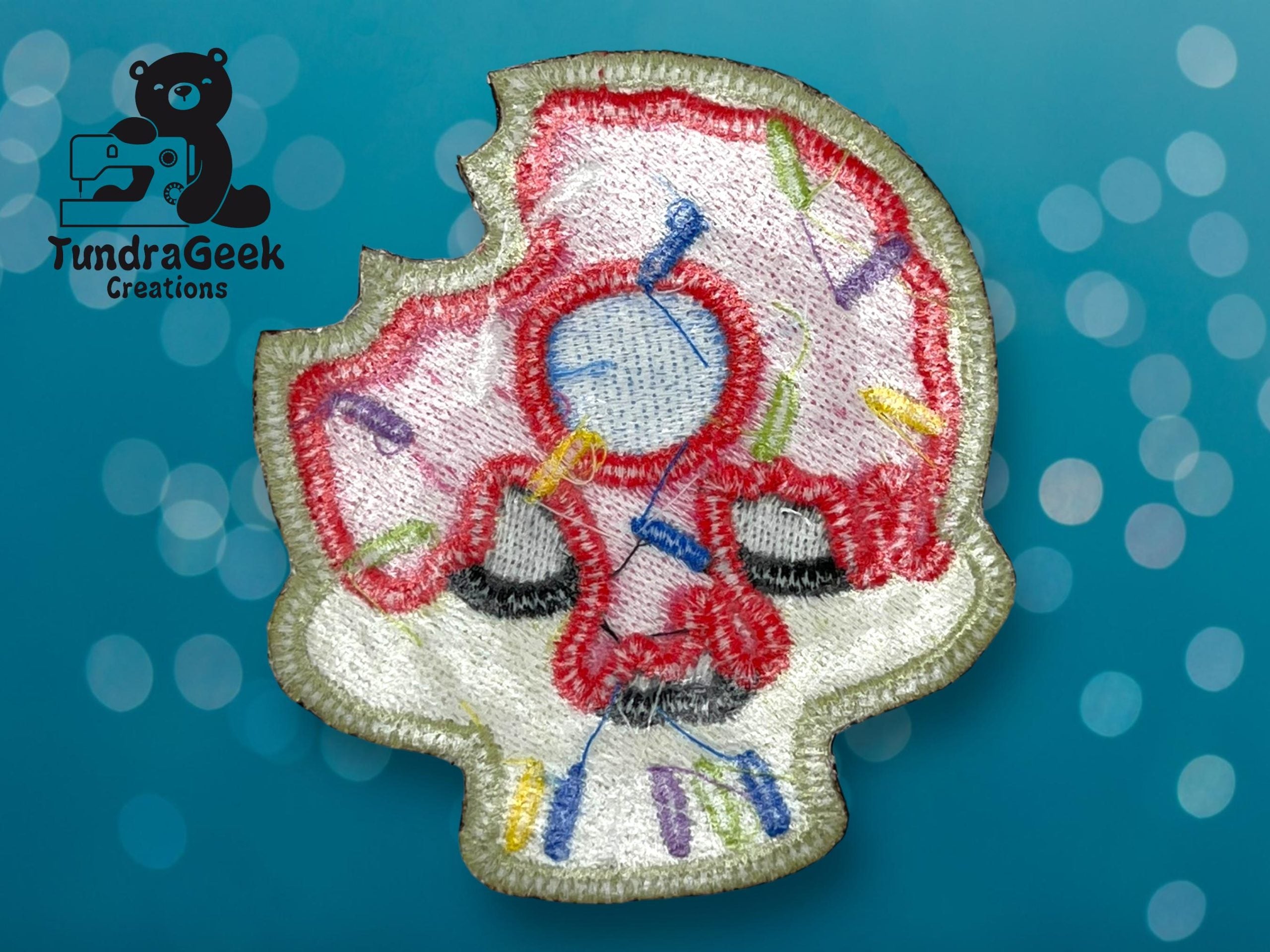 An embroidered patch shaped like a bitten skull with colorful thread accents is shown on a blue background. TundraGeek Creations logo appears in the top left corner.