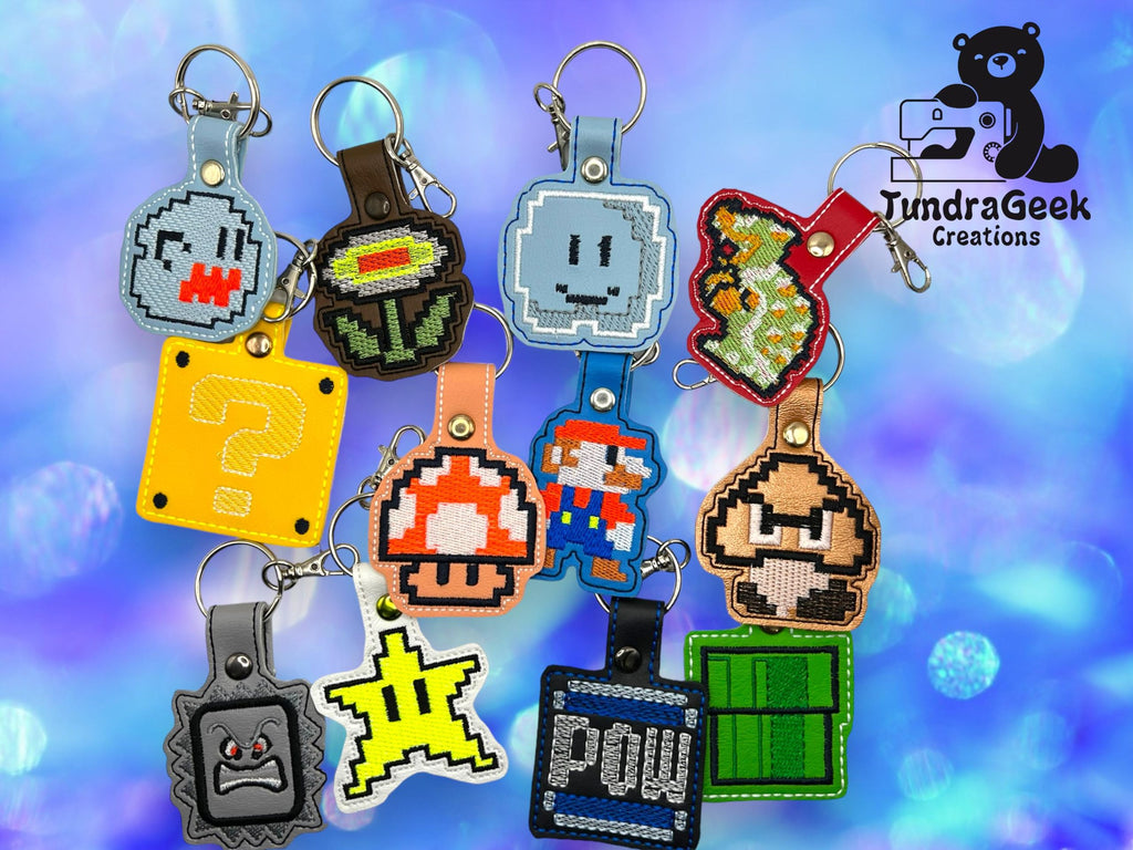A collection of keychains featuring pixel art designs inspired by classic Mario game characters and items, displayed on a blue bokeh background with the TundraGeek Creations logo.