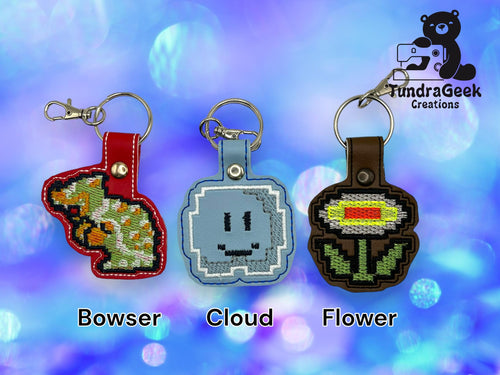Three embroidered keychains labeled Bowser, Cloud, and Flower are displayed on a blue bokeh background with the TundraGeek Creations logo in the top right corner.