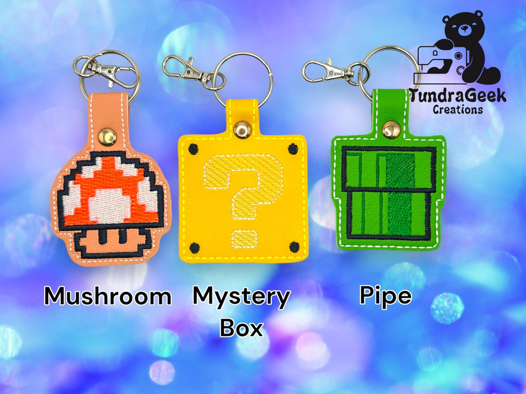 Three embroidered keychains shaped as a red mushroom, a yellow mystery box, and a green pipe are displayed on a blue bokeh background with TundraGeek Creations logo.
