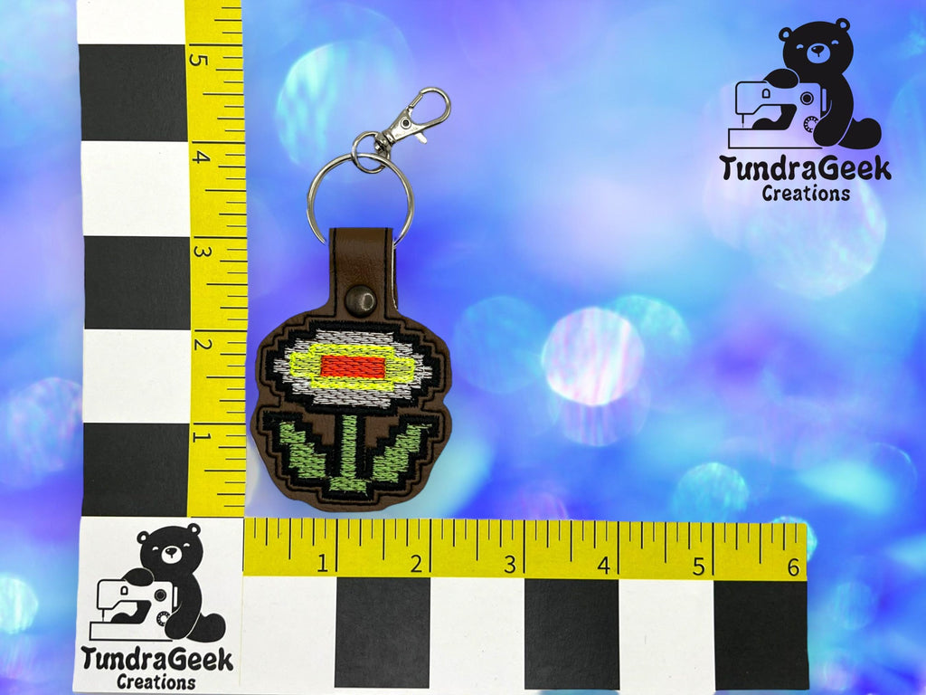 A keychain with a pixelated flower design is placed on a measuring mat with rulers, featuring the TundraGeek Creations logo on a blue bokeh background.