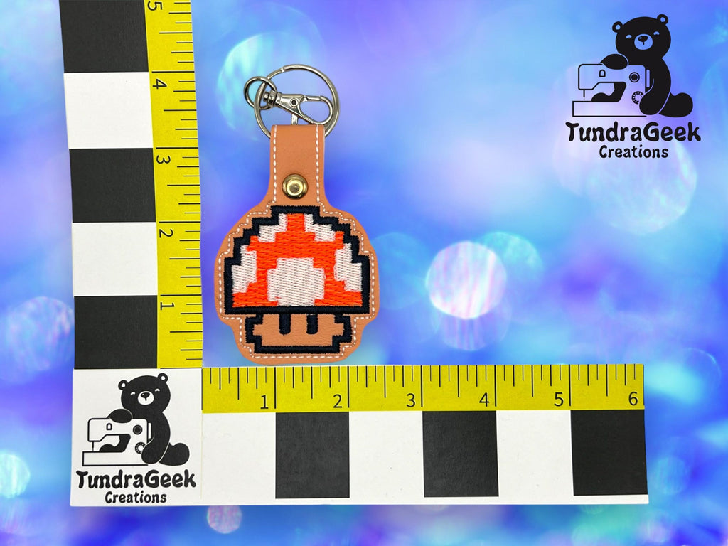 A pixelated mushroom keychain is displayed on a brown leather backing next to a ruler for scale, with a TundraGeek Creations logo in the corners on a blue background.