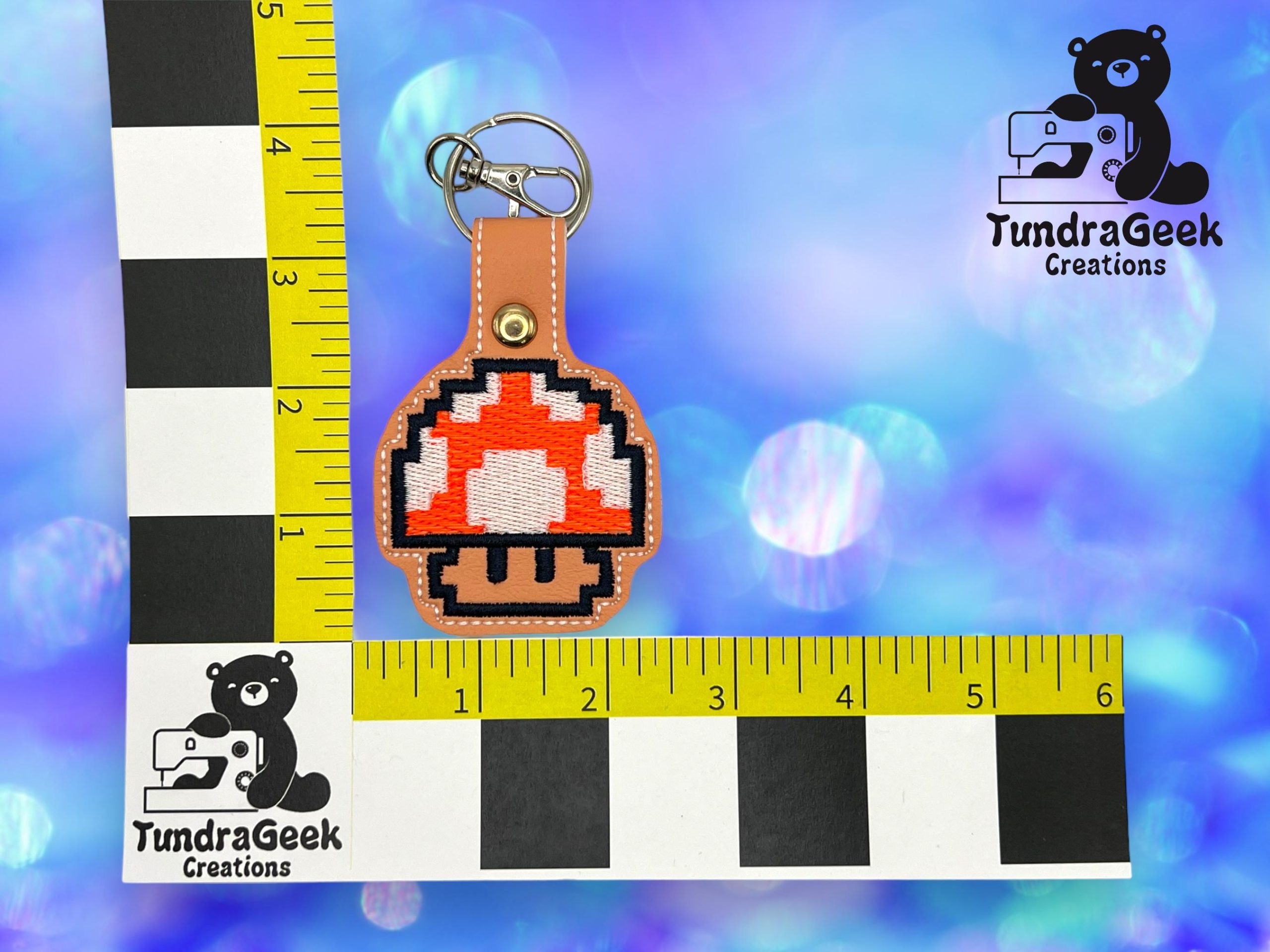 A pixelated mushroom keychain is displayed on a brown leather backing next to a ruler for scale, with a TundraGeek Creations logo in the corners on a blue background.