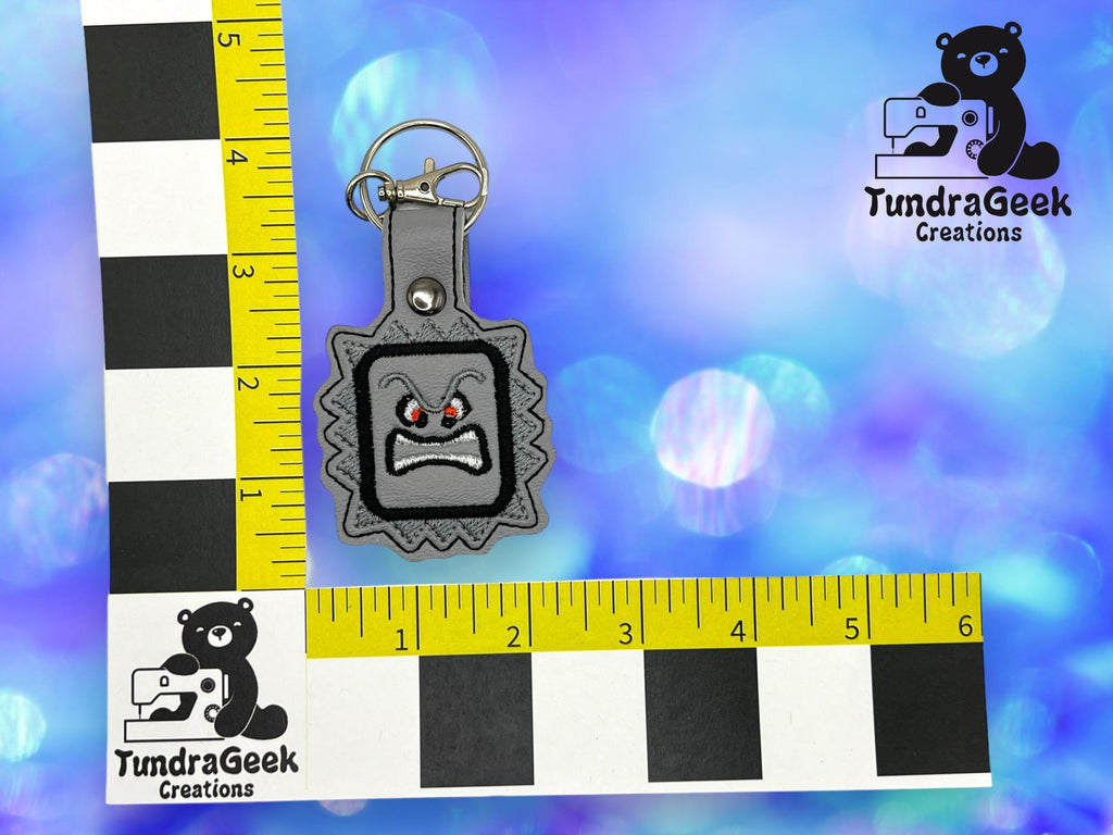 A gray keychain with a cartoon angry face is displayed on a blue bokeh background, next to two yellow and black TundraGeek Creations rulers for scale.