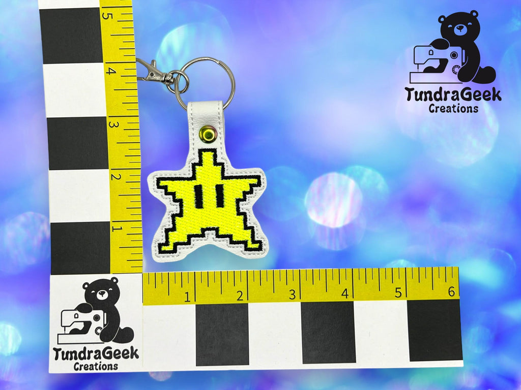 A keychain with a yellow pixelated star design is placed next to a ruler for scale, with the TundraGeek Creations logo in the background.