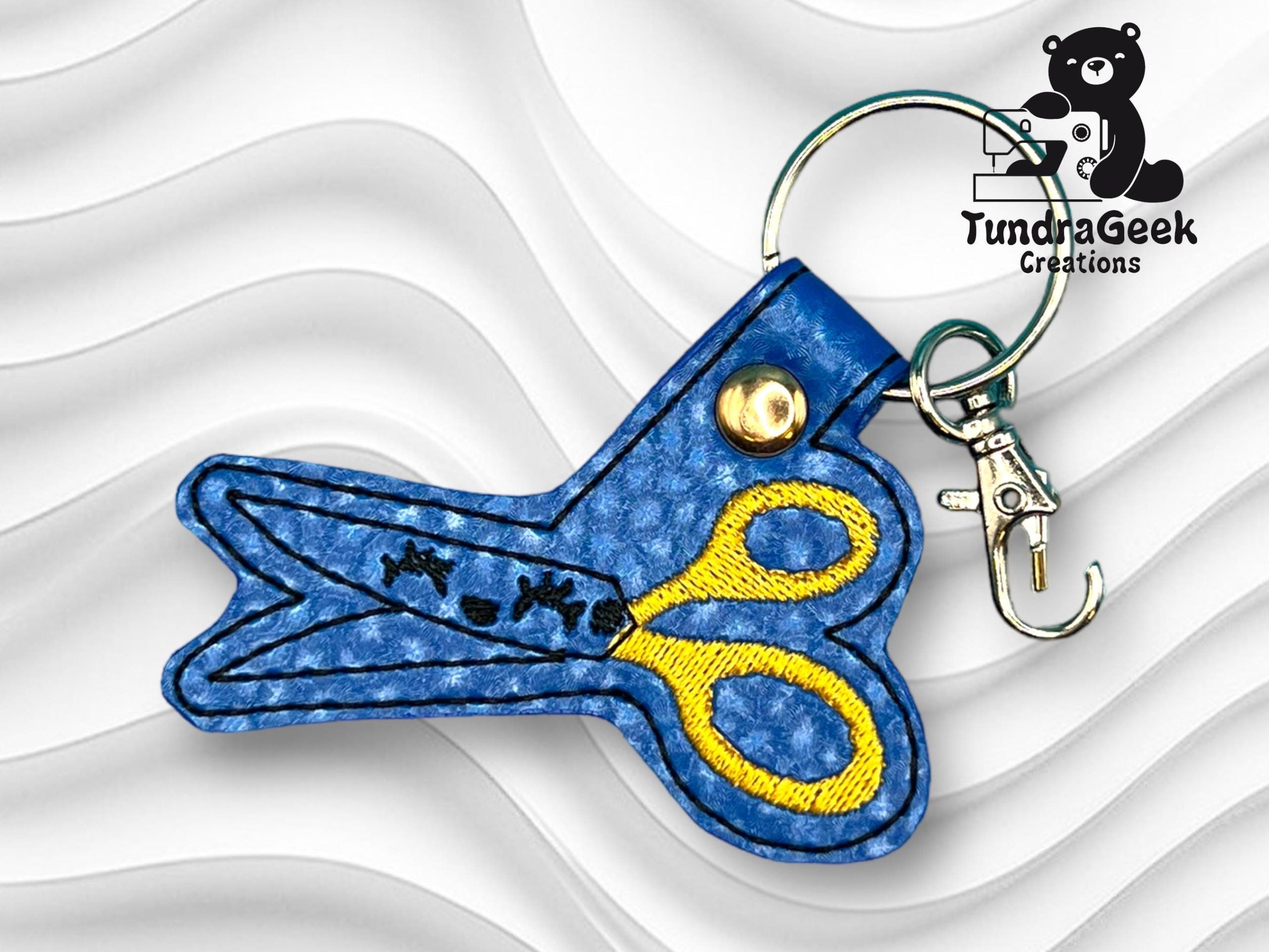 A blue leather keychain shaped like scissors with yellow and black stitching, metal clasp, and keyring; TundraGeek Creations logo in the top right corner.